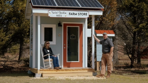 A small farm store with a sign reading 'Sweet Jordan Bees Farm Store' on its roof, featuring solar panels. A woman sits on a white rocking chair on the porch, while a man stands to the right, smiling and gesturing towards the store. The building is surrounded by trees, with a grassy area in front.