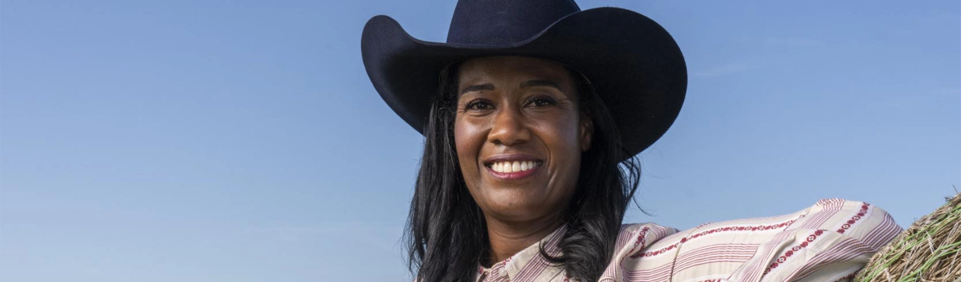 A smiling woman wearing a black cowboy hat and a striped Western-style shirt, standing outdoors against a clear blue sky with a bale of hay visible in the background.