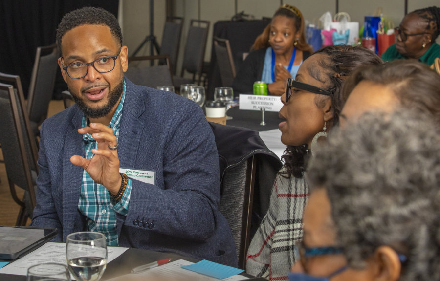 A man wearing glasses and a blue plaid shirt under a dark blazer gestures while speaking during a discussion at a conference table. He is engaged with several people around the table, including a woman in a plaid sweater and others listening attentively. Papers, glasses of water, and a nameplate are on the table, and more participants are visible in the background.