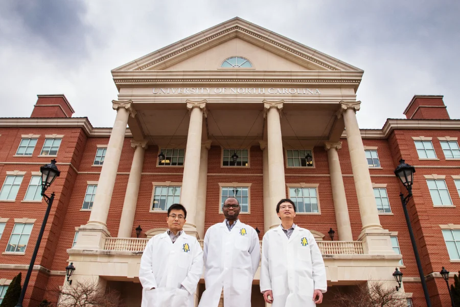 Three researchers wearing white lab coats stand in front of a red-brick building with large columns and a sign reading "University of North Carolina" under a cloudy sky.