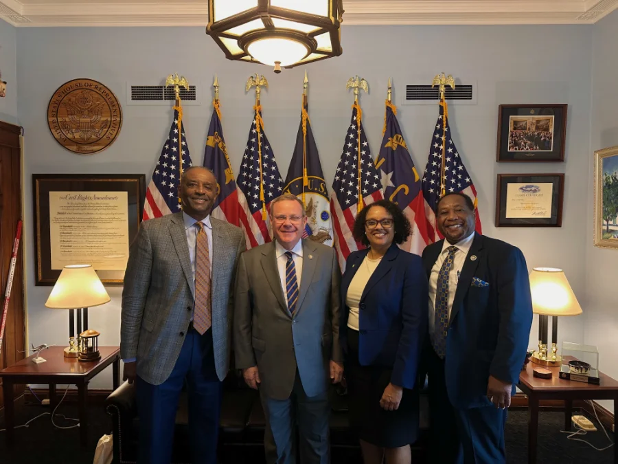 Four people stand together in a formal office setting, posing for a photo. Behind them are several U.S. and state flags, along with a seal that reads “U.S. House of Representatives.” The room has framed documents and photographs on the walls, two lit table lamps, and a large hanging light fixture overhead.